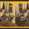 People on a porch -- two men playing chess accompanied by two women.