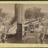 Street view including buildings with awnings across from a park, trolleys, carriages.
