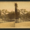 View of a park with a fountain in a lake and people on a bench in the background.