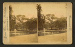 View of a park with a fountain in a lake and people on a bench in the background.