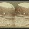 Man in cable car over shallow river.