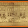 Boys and girls pose with adults on front steps of large brick institutional building.