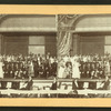 Performers in costume posing on stage with musicians in pit, viewed across orchestra seating.