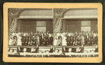 Performers in costume posing on stage with musicians in pit, viewed across orchestra seating.