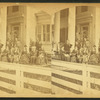Multi-generation male and female family members, wearing suits and dark dresses, pose before white-columned porch and trellis of multi-story clapboard house; white board fence in foreground. 