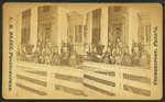 Multi-generation male and female family members, wearing suits and dark dresses, pose before white-columned porch and trellis of multi-story clapboard house; white board fence in foreground. 