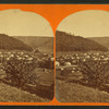 Small village at foot of hill; small tree and railroad car in foreground.