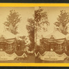 Decorated tent [Fourth of July?], with American flags, ornamental plants, liberty bell in flowers, and drapery bearing phrase 'Virtue Liberty & Independence;' bearded, hatted man stands inside.]