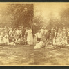 Large group of men, women and children on a lawn under some trees.