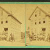 Men in front of four story clapboard building with wagons.
