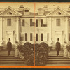 Man in tophat and woman wearing dress with bustle and train, in front of large clapboard house.