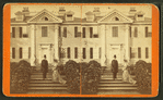 Man in tophat and woman wearing dress with bustle and train, in front of large clapboard house.