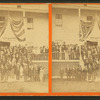 Group of men and women posing on steps of large building, flag draped overhead, and a sign reading "Headquarters First ME Cavalry".
