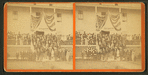 Group of men and women posing on steps of large building, flag draped overhead, and a sign reading "Headquarters First ME Cavalry".
