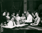 Stephen Sondheim on piano and Leonard Bernstein standing amongst female singers rehearsing for West Side Story (variant).