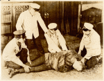 Harry Houdini on floor in straight jacket, surrounded by four unidentified men in white jackets and caps