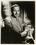 Portrait of Tennessee Williams sitting in chair with hands clasped holding a cigarette holder.