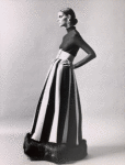 Unidentified model wearing dark Norman Norell evening dress with wide belt, dark top with turtleneck and floor length skirt with fur ruff