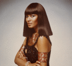 Head and shoulders shot of model wearing straight darkshoulder-length Egyptian style wig beaded dress with Egyptian broach between breats red and gold armbands and lion collar