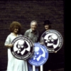 Actors(L-R) Cleo Laine, George Rose and Betty Buckley holding large "Free Shakespeare in the Park" signs while performing in the New York Shakespeare Festival's production of the musical "The Mystery of Edwin Drood" at the Delacorte Theatre in Central Park. (New York)