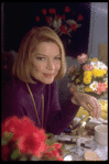 Actress Ellen Burstyn sitting at vanity table between 2 bouquets of flowers and toying with a small mirror