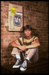 Publicity photo of actor Fisher Stevens sitting in front of poster for Broadway play "Brighton Beach Memoirs" (New York)