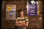 Publicity photo of actor Fisher Stevens standing in front of posters for Broadway play "Brighton Beach Memoirs" & "Torch Song Trilogy", both of which he appeared in (New York)