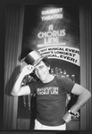 Theatrical producer Joseph Papp in "A Chorus Line" t-shirt, posing with a golden top hat in front of poster proclaiming "A Chorus Line" as Broadway's longest running show.