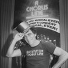 Theatrical producer Joseph Papp in "A Chorus Line" t-shirt, posing with a golden top hat in front of poster proclaiming "A Chorus Line" as Broadway's longest running show.