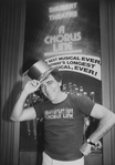 Theatrical producer Joseph Papp in "A Chorus Line" t-shirt, posing with a golden top hat in front of poster proclaiming "A Chorus Line" as Broadway's longest running show.