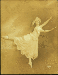 Dancer in arabesque, wearing large hat and long scalloped-edge skirt